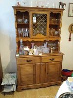 Front view of RAD wooden china cabinet with hutch showing glass door, open shelves, two drawers and two cabinet doors at the bottom with various glassware displayed on top.
