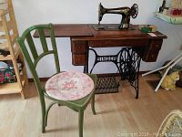 Singer treadle sewing machine mounted in oak cabinet with drawers. Old wooden green painted chair with floral round seat cushion placed beside the cabinet.