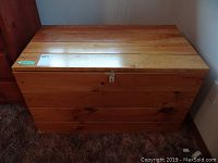 Front view of pine wood chest showing natural wood grain and padlock hasp, resting on carpet.