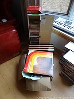 Wide view of vinyl records in cardboard box with grainy album covers and vertical beige CD storage stand.
