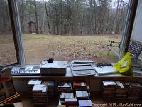 Wide shot showing from left to right an Equip keyboard, Canon printer, Brother electronic typewriter, Zenith DVD player, and a yellow plastic bag on a windowsill; several media cases stacked below the window ledge.