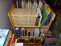 Front view of the small wooden bookshelf filled with books including a set of books with white spines, a biography of Bill Clinton, and various others.