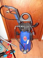 Front view of blue Simoniz pressure washer with black hose reel and coiled hose, black wand with trigger handle attached to top, showing wear and use.