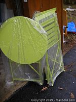 Lime green metal round folding table and two matching folding chairs in clear plastic packaging, leaning against wall.