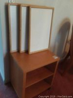 Wooden rolling cart with two open shelves and three wooden bed trays with white surface and wooden frame leaning against wall on top of cart