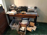 Office desk with three printers, computer keyboards, telephones, boxes with cables and office supplies beneath desk