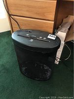 Black electric paper shredder in front of a wooden desk, showing the main unit with a bin base for shredded paper collection.