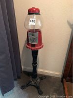 Full height image of freestanding vintage gumball machine showing red body, glass globe top, and cast iron base on carpet.