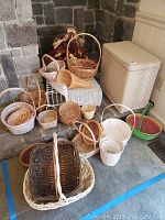 Full view of assortment of 21 baskets arranged in front of stone wall and beside white wicker hamper with lid.
