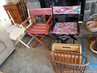 Three director chairs and two wood magazine racks arranged on floor including a white frame chair with white fabric seat/back, a natural wood frame chair with red striped fabric, a black frame chair with colorful patterned fabric, and two wood magazine racks (rectangular with slats and oval basket style).