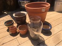 Photo showing a group of various sized terra cotta and clay pots along with a large clear glass vase on wooden surface outdoors.
