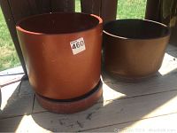 Two outdoor round ceramic pots in reddish-brown with matching saucers. Larger pot in foreground and smaller pot behind, placed on wooden surface outdoors.
