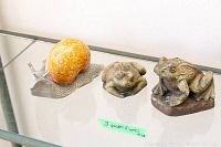 Three garden ornaments: metal and resin snail, small resin frog, larger resin toad, displayed on a glass shelf