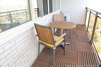 View of the round table and two outdoor chairs on balcony showing wood slat design and metal frame construction, yellow seat cushions on chairs visible.
