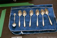 Six sterling silver espresso spoons and one souvenir spoon laid out on blue felt cloth, showing the front side with polished bowls and decorated handles.