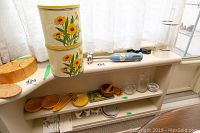 Cuisinart Smart Stick hand blender shown with other kitchen ware on windowsill including vintage tin canisters with sunflower design, wooden mortar and pestle, glass cups, and assorted kitchen dishes.