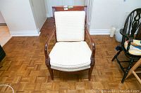 Front view of vintage-style carved mahogany chair with light cream textured upholstery and visible parquet floor.