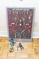 Overall view of framed wine opener plaque leaning against wall with assorted openers on floor