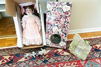 Porcelain doll laying inside original floral box with stand and 3 small white china elephant figurines placed in front alongside a floral resin photo frame on a patterned rug.