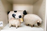 Shelf display of three farm animal figurines: a black and white cow and two sheep figurines, one larger white and one smaller cream colored.