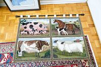 Full view of the needlepoint farm animals tapestry lying flat on the floor next to framed art and a rug, showing its overall size, design, and hanging rod attached.