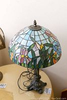 Front and angled view of the Tiffany style stained glass lamp with colorful floral patterns on the shade and a metal female figure base on a table.