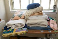 Wide view of all linens stacked on table near window, showing assorted quilts, blankets, towels, and pillows.