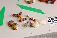 Photo showing all six pairs of pierced earrings spread on a surface with green tape background, including round metal and wood tone, amber and black beads, cream shell-shaped dangles, silver metal and pearl drops, and red stone drops.