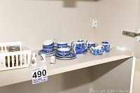 Blue willow style children's tea set arranged on shelf including teapot, sugar bowl, five cups and saucers, and plates with three decorative thimbles and two napkin rings nearby.