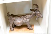 Front-left view of a heavy metallized carved goat sculpture showing intricate detailing and mounted on a wooden base.
