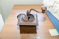 Front angle view of the antique coffee grinder on wooden table showing the cast iron body, small drawer at the front, and wooden crank handle.