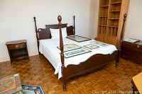 Gibbard mahogany queen four poster bed shown from side angle with mattress and white bedding, small bedside table near wall. Several framed artworks placed on mattress.