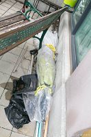 Outdoor umbrella wrapped in clear plastic and tied with yellow ropes laid on ground near bench.