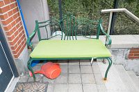 Full view of the green metal outdoor bench with green cushion seat placed outdoors on tiled ground next to a brick wall and greenery.