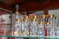 Gold trim glassware set including tall beer glasses, wine glasses, sherry glasses, and a decanter with stopper, all arranged in front of a mirrored wood cabinet.
