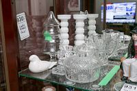 Nine pieces of assorted glassware including different styles of clear glass candle holders, a cut glass covered bowl, a glass pitcher, a glass tree-shaped cover, and a white ceramic bird figurine on a glass shelf.