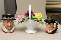 Two mini Toby Royal Doulton character mugs (Beefeater and Night Watchman) and one white ceramic basket with colorful ceramic flowers arranged on a beige surface.