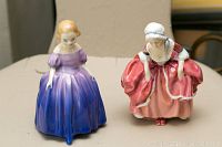 Photo of two mini Royal Doulton figurines, Goody Two Shoes (purple dress) and Marie (red dress) showing front and side angles on a table.