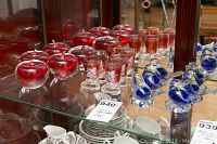 Red glass apple figurines, blue glass bird figurines, and vintage boot-shaped shot glasses displayed on glass shelving.
