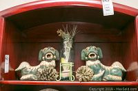 Pair of green pottery foo dogs on a wooden shelf with glass vase and decorative pine cones.