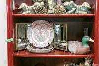 Display cabinet shelf with pottery duck on right, three decorative plates in center front, and folding three-frame vintage mirror behind plates.