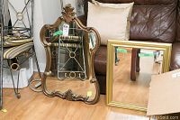 Both mirrors shown next to chair and couch on floor, one ornate wooden framed mirror with scrolling decorations, one square gold framed mirror leaning against couch