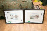 Photo showing two framed prints leaning against furniture on floor, both with rural house scenes, framed with dark wood frames and double mats