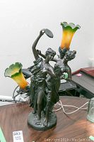 Metal figurine table lamp with two female dancers and two flower-shaped art glass lampshades, yellow-green gradient, shown on a wooden surface with a bicycle in background.