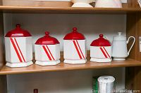 Four white china canisters with red lids and diagonal red stripes on a wooden shelf.