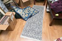 Two area rugs on a wood floor - a smaller blue floral rug partially folded and a longer gray and white patterned runner rug flat.