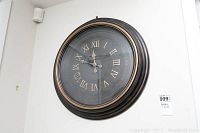 Front angled view of clock mounted on wall, showing black and bronze frame and Roman numeral face.