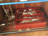 Wooden box filled with assorted Wallace stainless steel flatware, two brass candlesticks, and a clear glass vase placed around it.