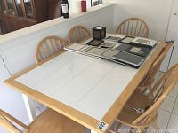 Rectangular kitchen table with white tile top and light wood frame with six matching spindle-back wooden chairs around it. The table surface has several picture frames and small decorative objects on top.