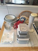 Photo of kitchen items on a table including plates, glass cups, red teapot, plastic storage containers, paper towel holder, linens and napkin holder.